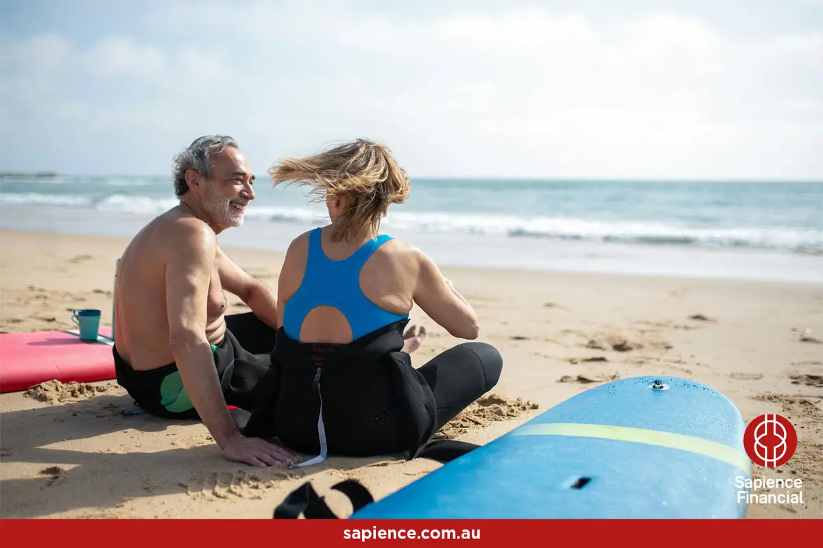 elder man and woman sitting on sand wearing wetsuits while having a conversation